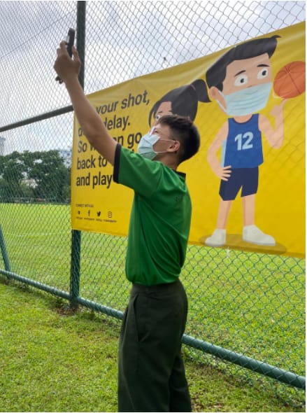 A masked young man in a green shirt takes a selfie in front of a yellow vaccination campaign banner on a fence.