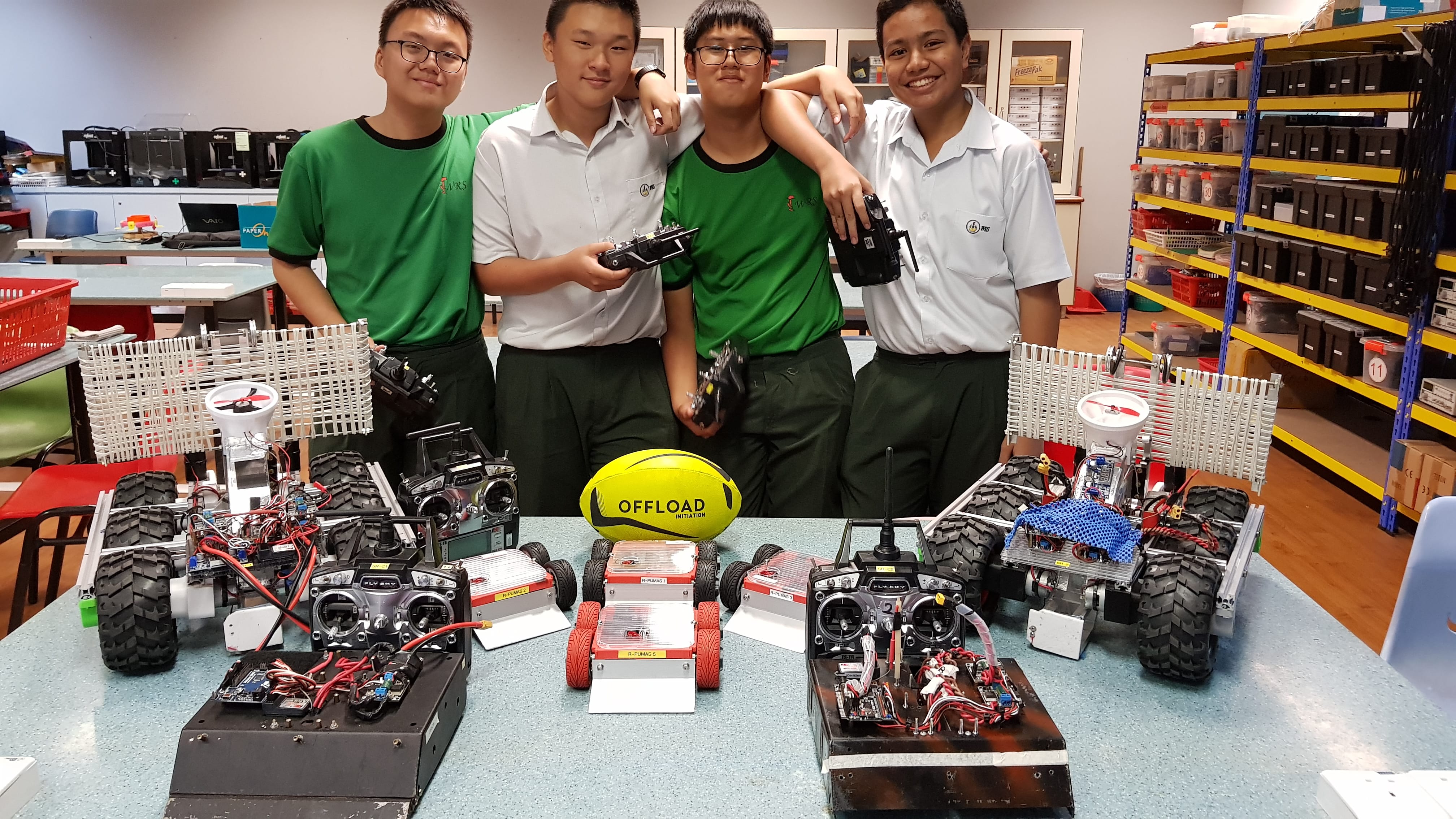 Four students in school uniforms stand behind a table displaying several built RC robots and a yellow rugby ball.