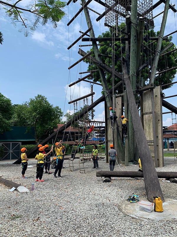 Children in yellow shirts and orange helmets climb and navigate a tall outdoor wooden rope course structure.