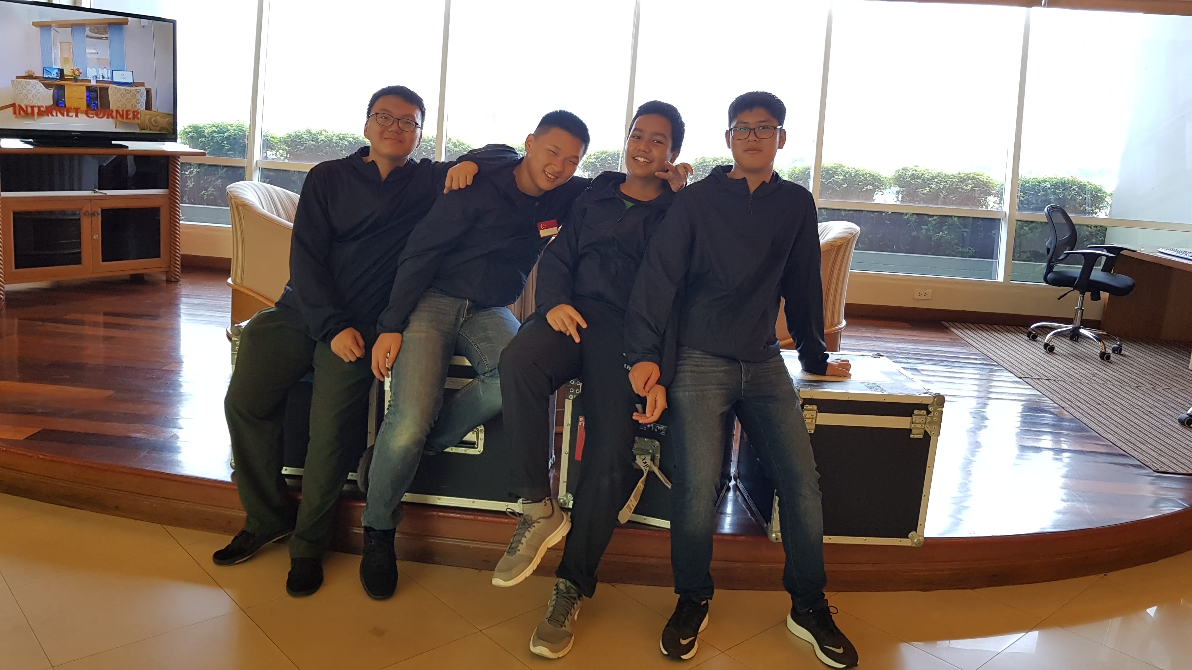 Four young men in dark jackets pose together sitting on a black equipment case in a bright indoor lobby.