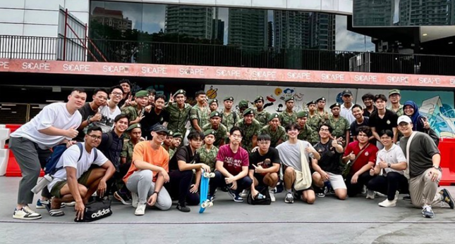 Large group of people, some in military camouflage uniforms, posing together outdoors in front of a Scape banner.