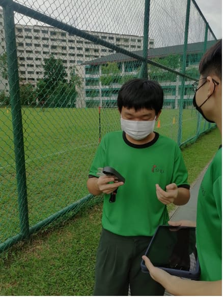Two masked students in green shirts stand near a green fence on a sports field, examining a small device.