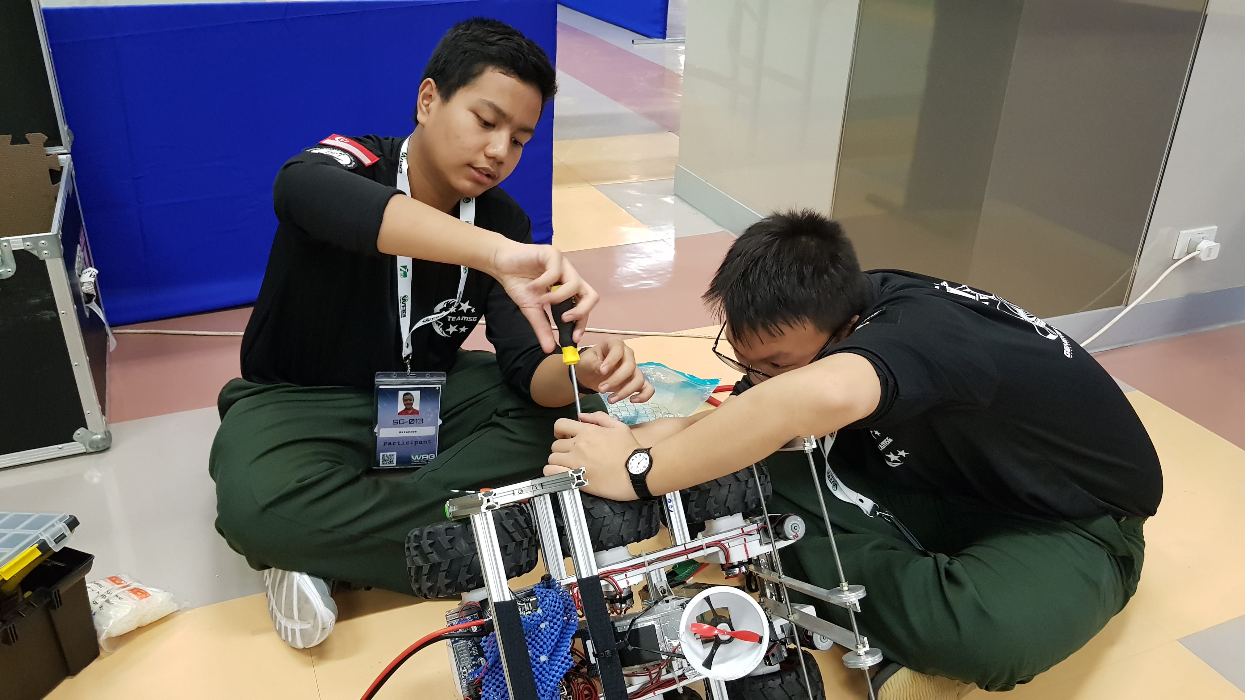 Two boys in black shirts sit on the floor assembling a wheeled robot, one using a screwdriver.