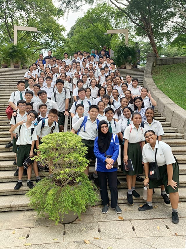A large group of uniformed students and a teacher pose together on outdoor stone steps surrounded by trees.