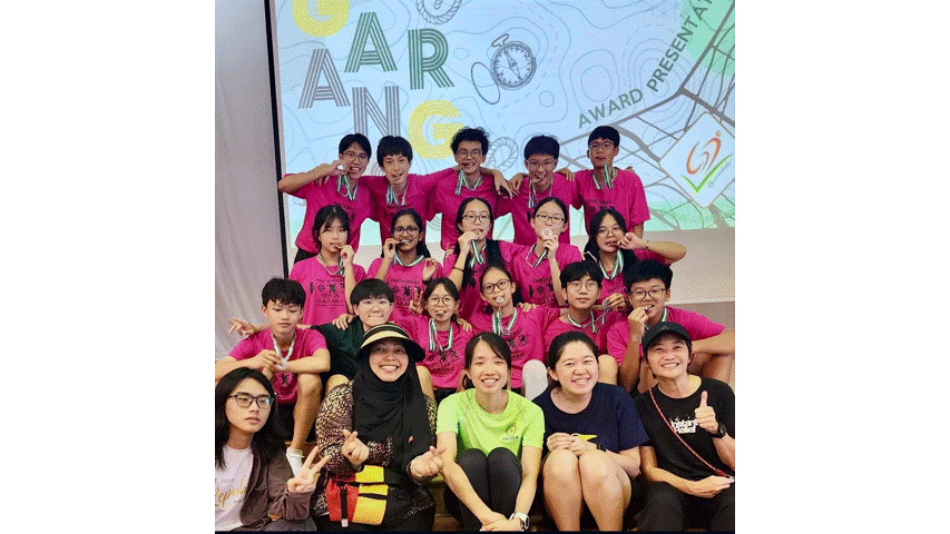 A group of students in pink shirts wearing medals pose together in front of an award presentation backdrop.