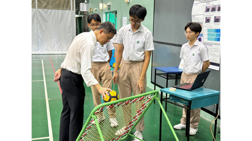 An adult in a white shirt bends to place a soccer ball into a green net frame while three students watch nearby.