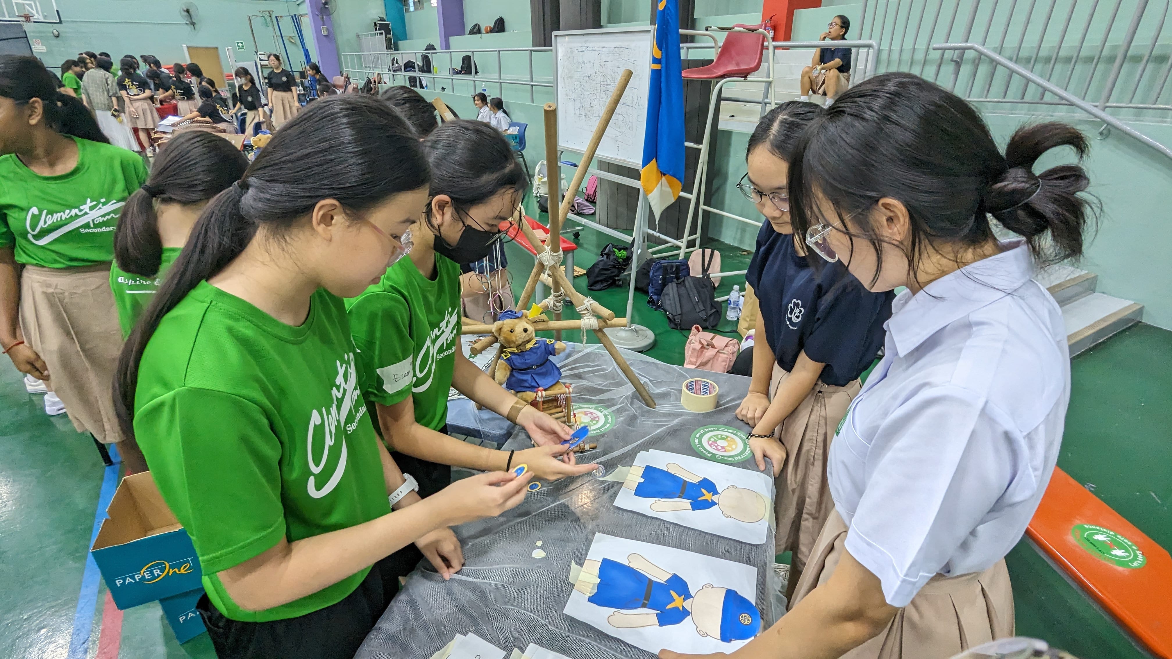 Students in green and school uniforms gather around a table with craft materials and a dressed teddy bear in a gymnasium.