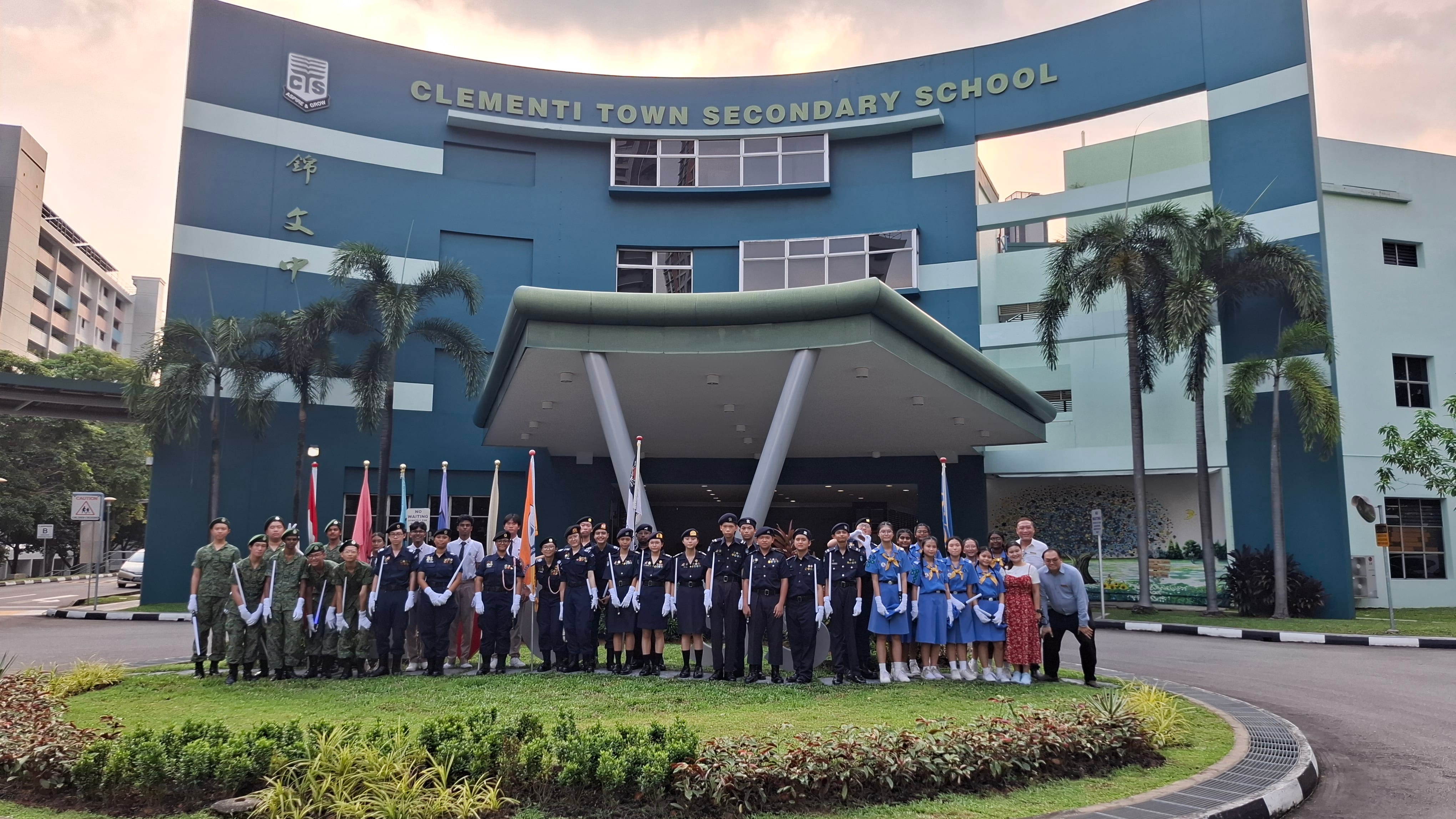 Students in uniforms holding flags stand in front of Clementi Town Secondary School's blue building entrance.