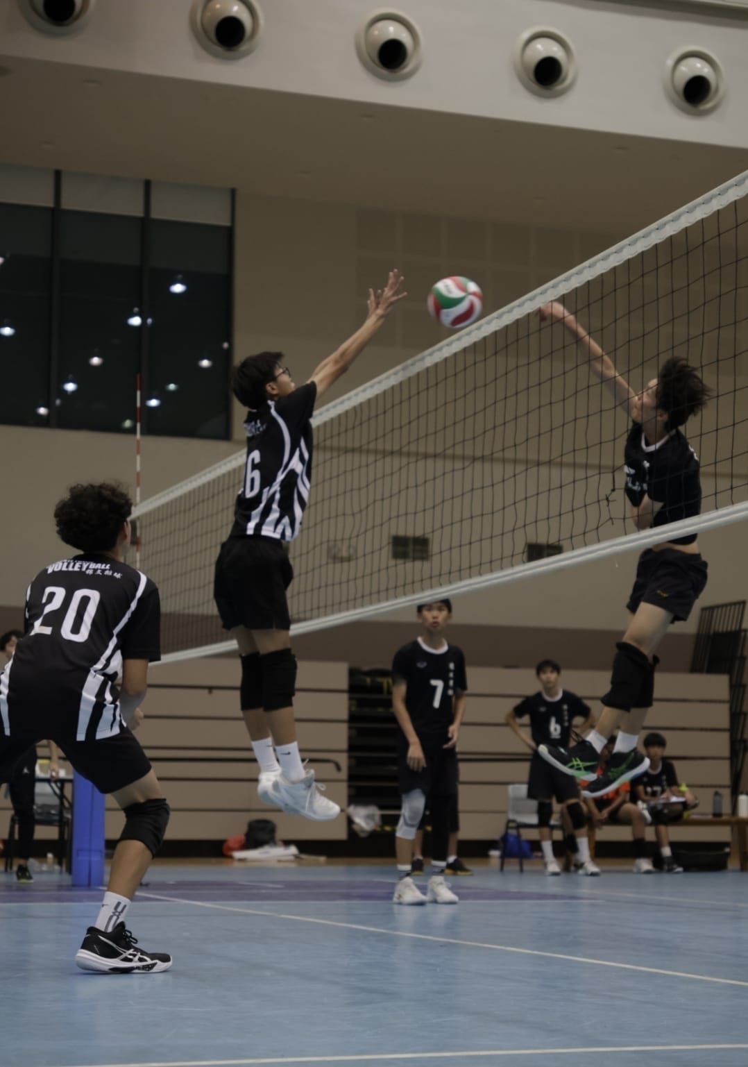 Two players in black uniforms jump at the net during an indoor volleyball match, with the ball in the air.