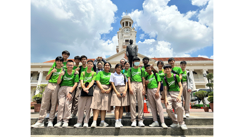 A group of students in green shirts pose on steps before a white clock tower building under a blue sky.