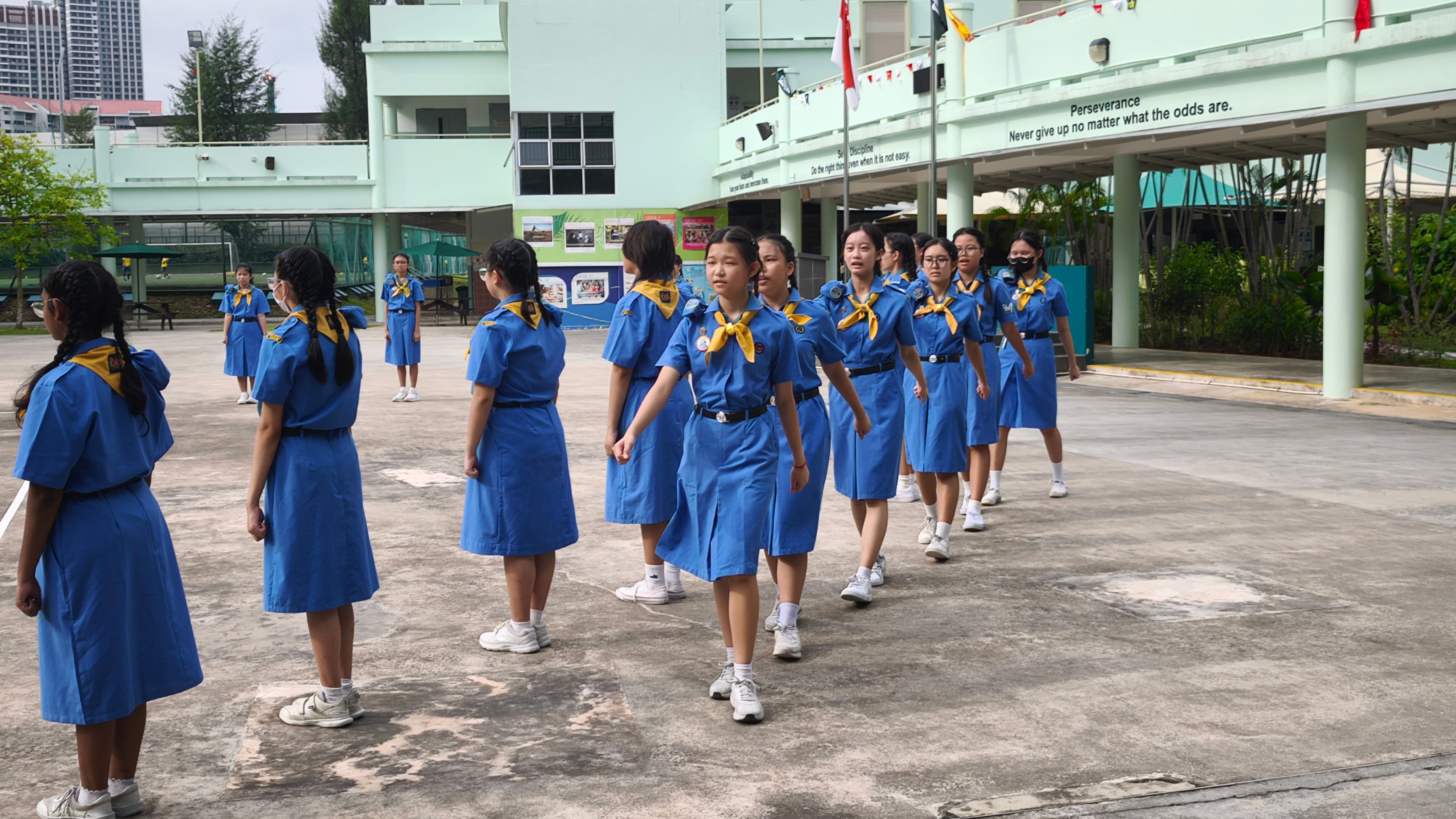 Girls in blue scout uniforms with yellow scarves marching in formation on a school courtyard.