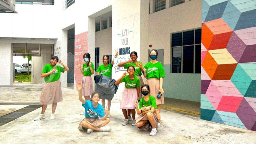 Eight students in green shirts pose outside a school building with colorful murals and a "Let Your Dreams" wall.