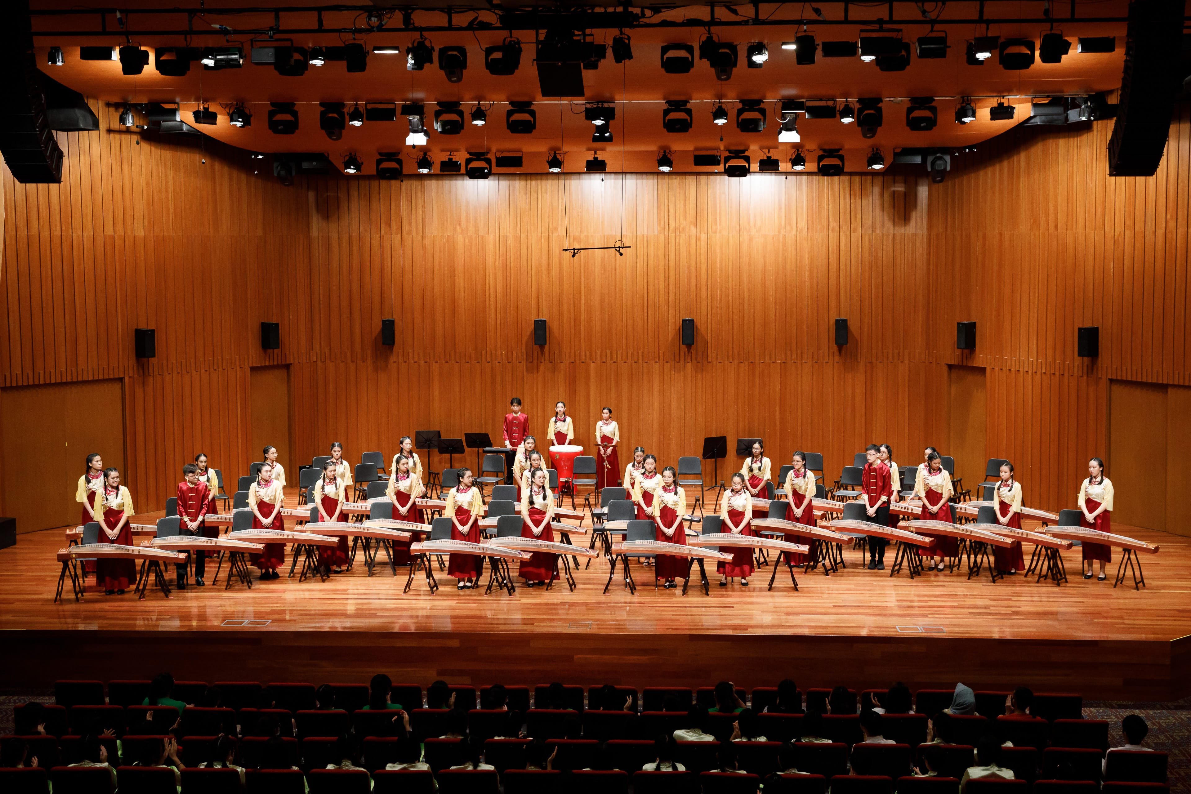 A large group of young performers in red and gold traditional outfits stand beside guzheng instruments on a concert stage.