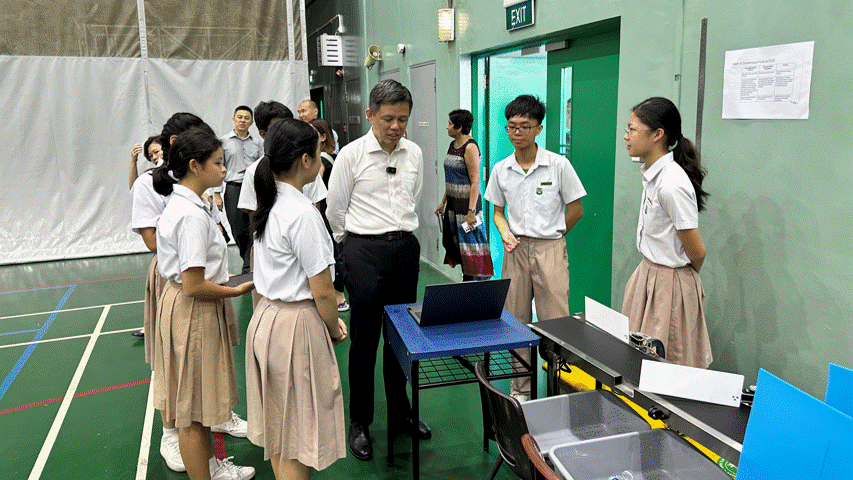 Students in school uniforms gather around a table with a laptop in a sports hall, speaking with an adult in a white shirt.