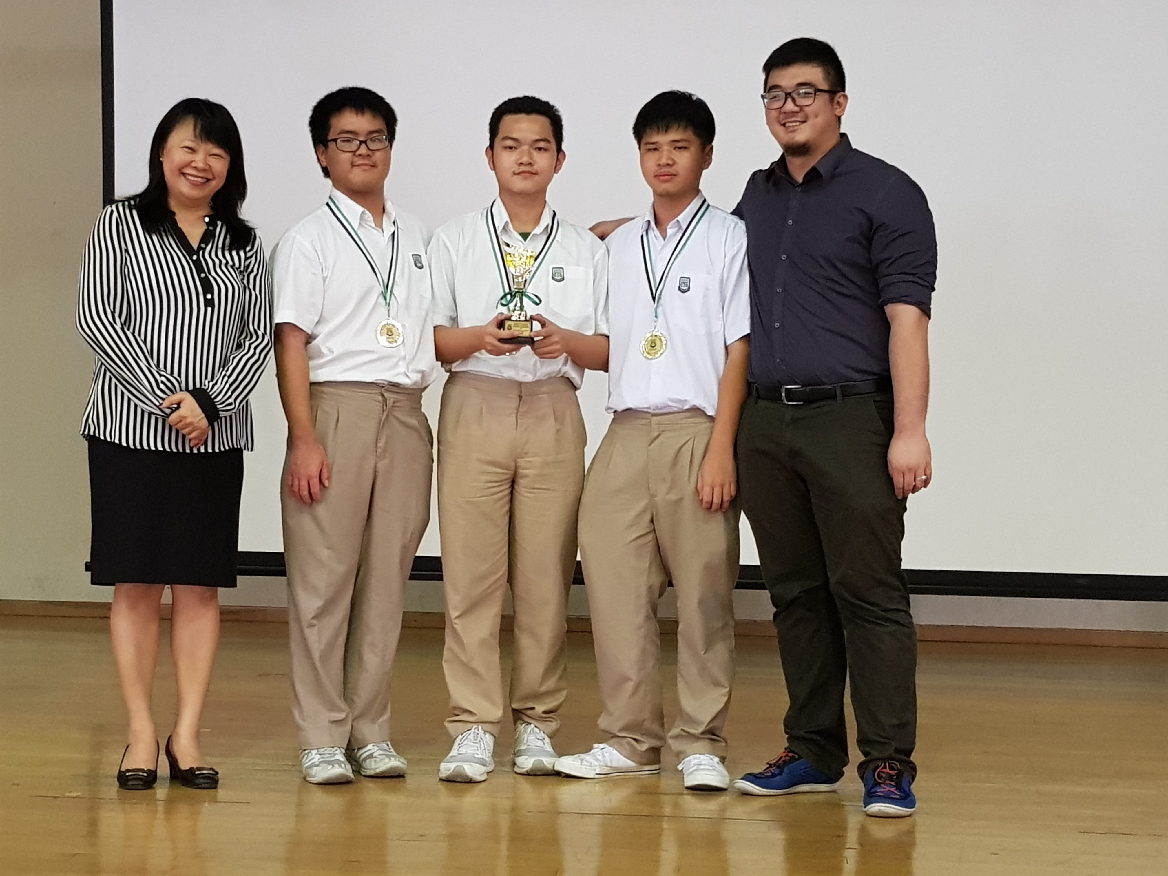 Five people pose together; three boys in school uniforms wear medals, one holds a small trophy.