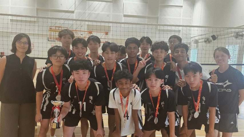 A youth volleyball team in black uniforms poses with medals and a trophy in front of a net indoors.