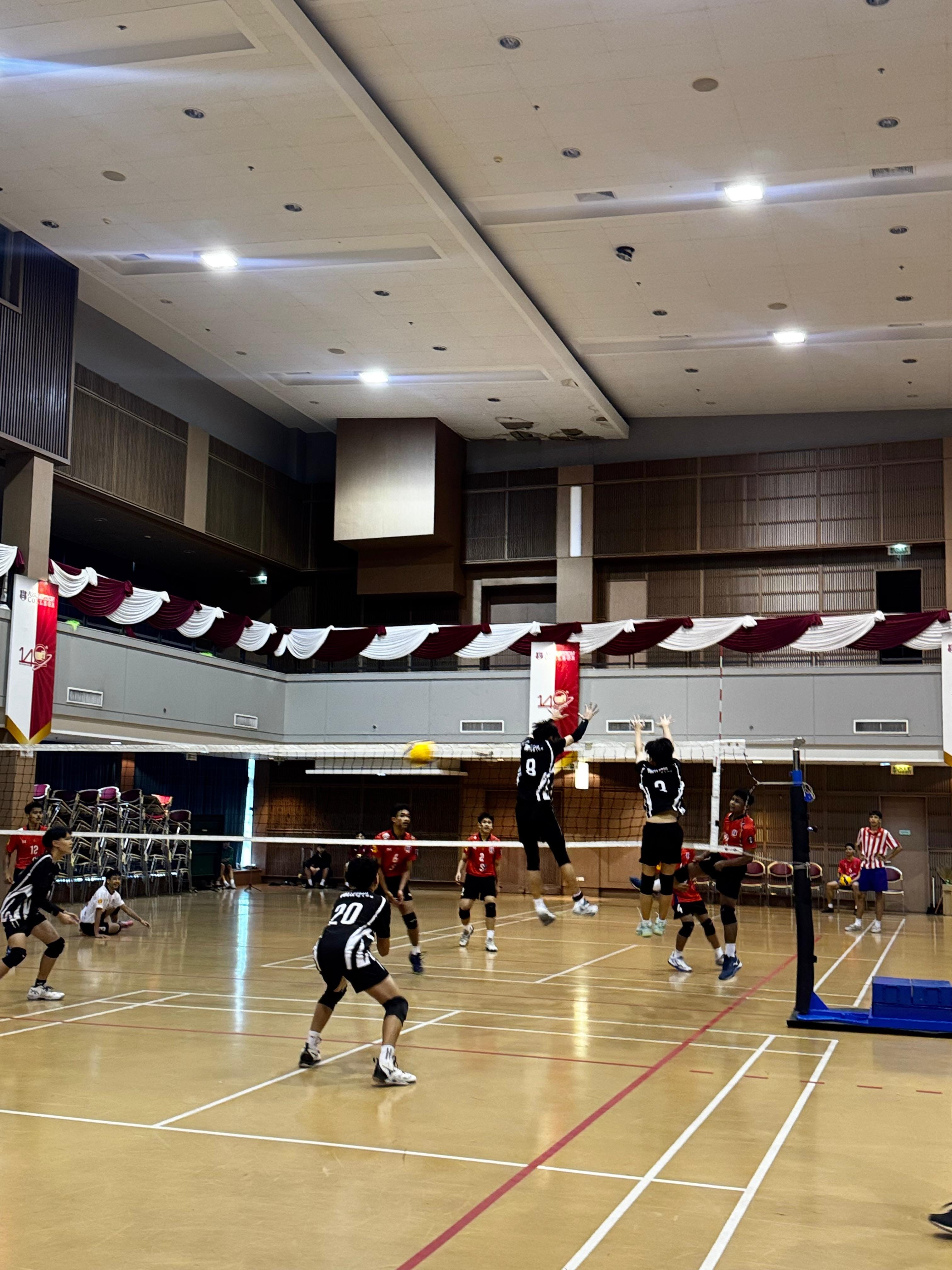 Two volleyball teams in black and red uniforms compete indoors, with players jumping at the net during a rally.