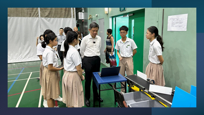An adult in a white shirt speaks with uniformed students gathered around a table in a school gym.