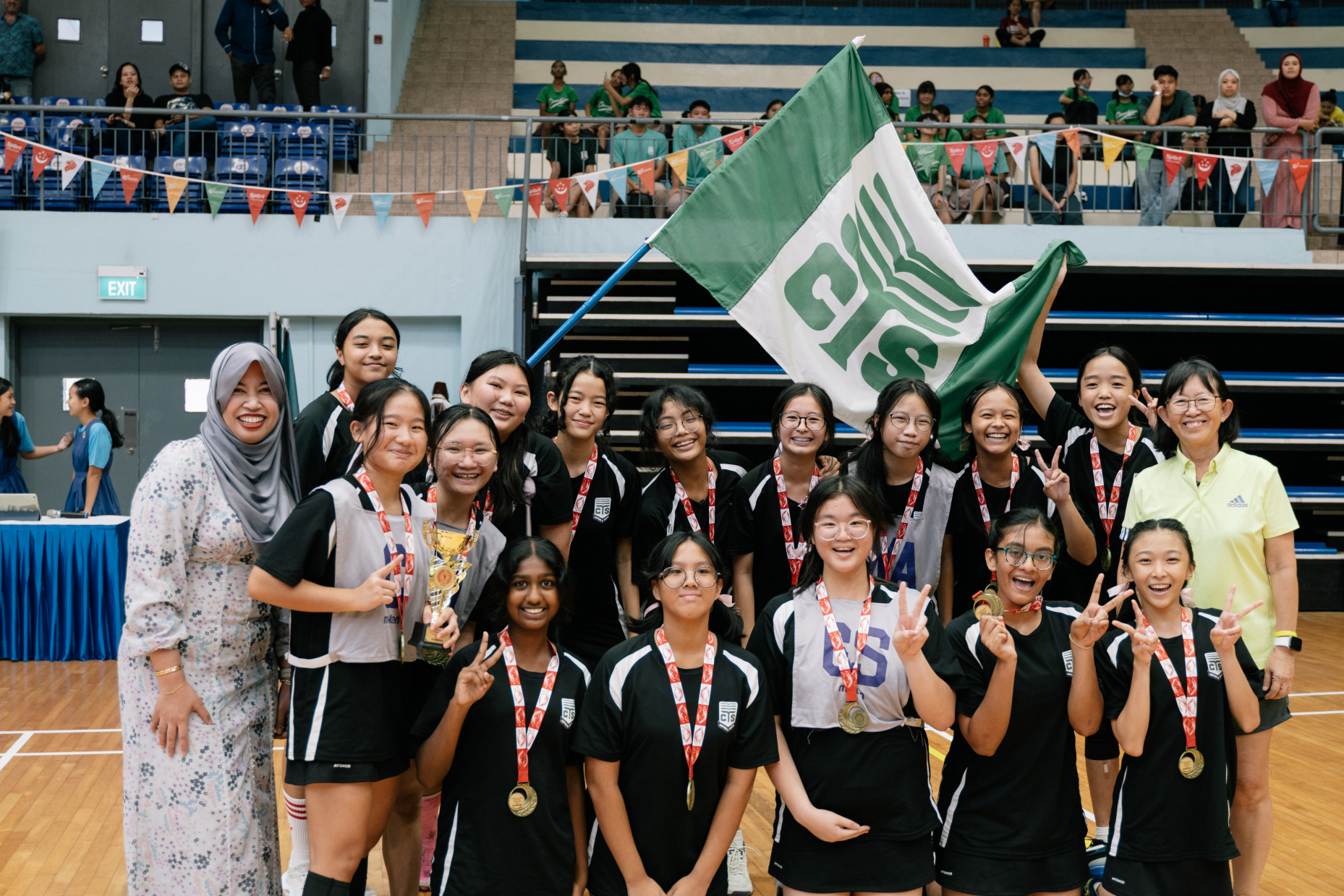Group of girls in sports uniforms with medals, holding a trophy and a green-and-white flag in a gym