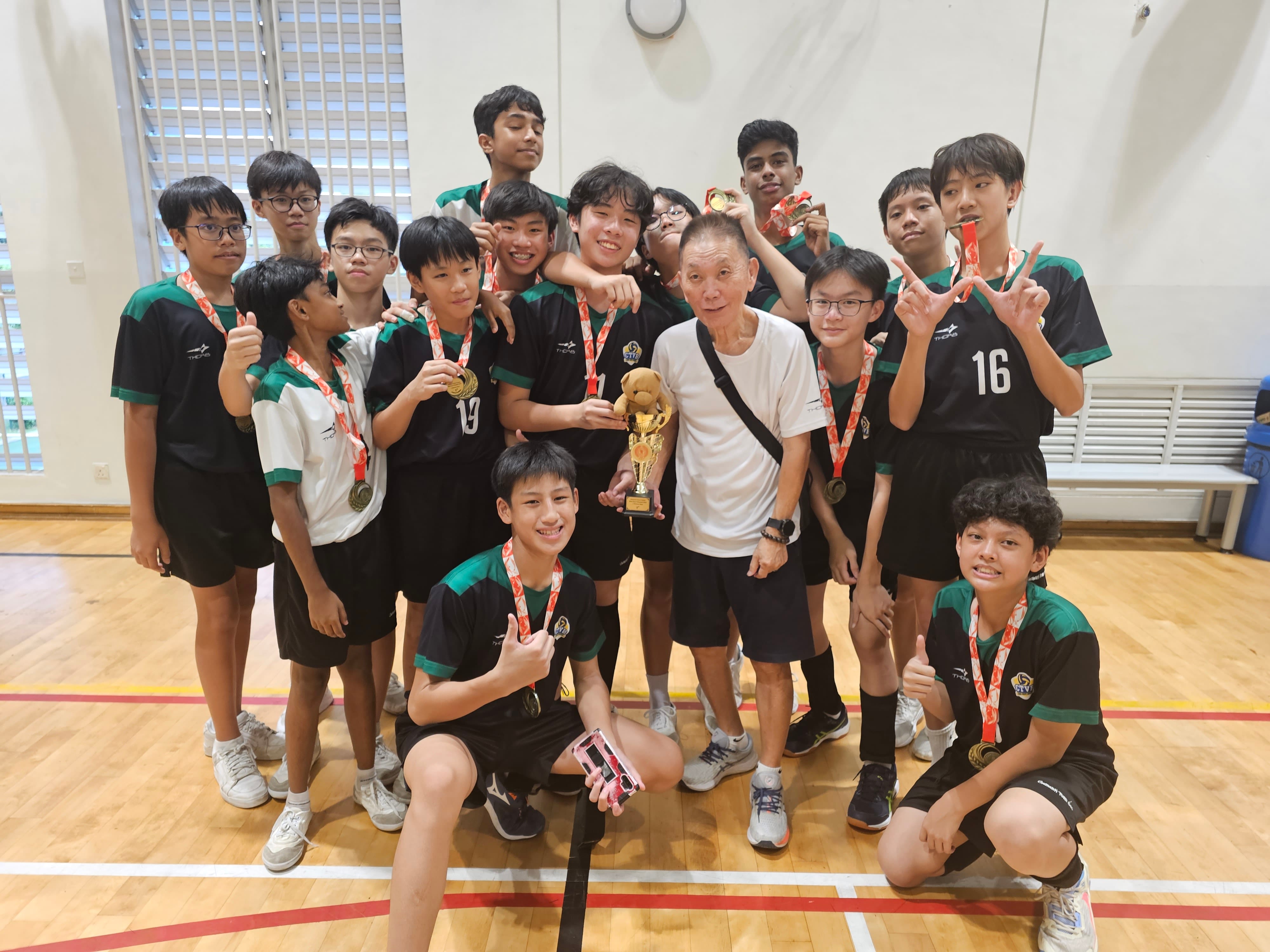 A youth sports team in black and green jerseys poses with medals and a trophy alongside their coach in a gymnasium.