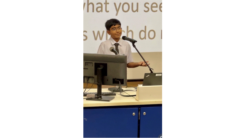 A young man in a white shirt and tie speaks at a microphone behind a podium with a monitor and projection screen behind him.