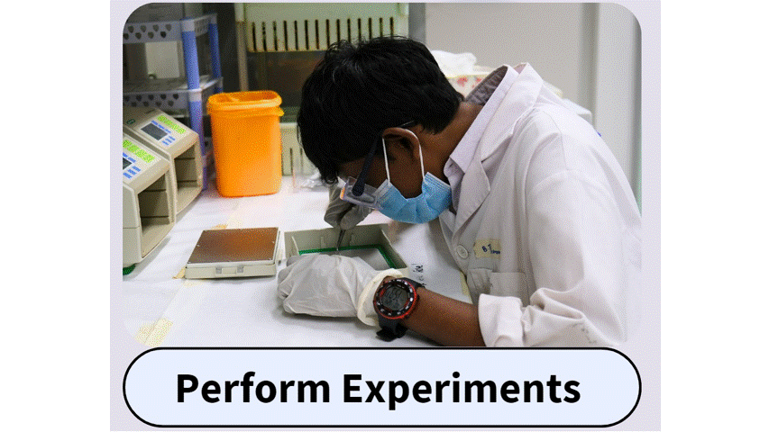 A lab worker in a white coat and face mask leans over a tray on a lab bench with equipment nearby.