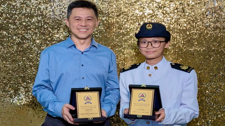 Two people smile while holding award plaques in front of a gold sequined backdrop; one wears a blue uniform with a cap.