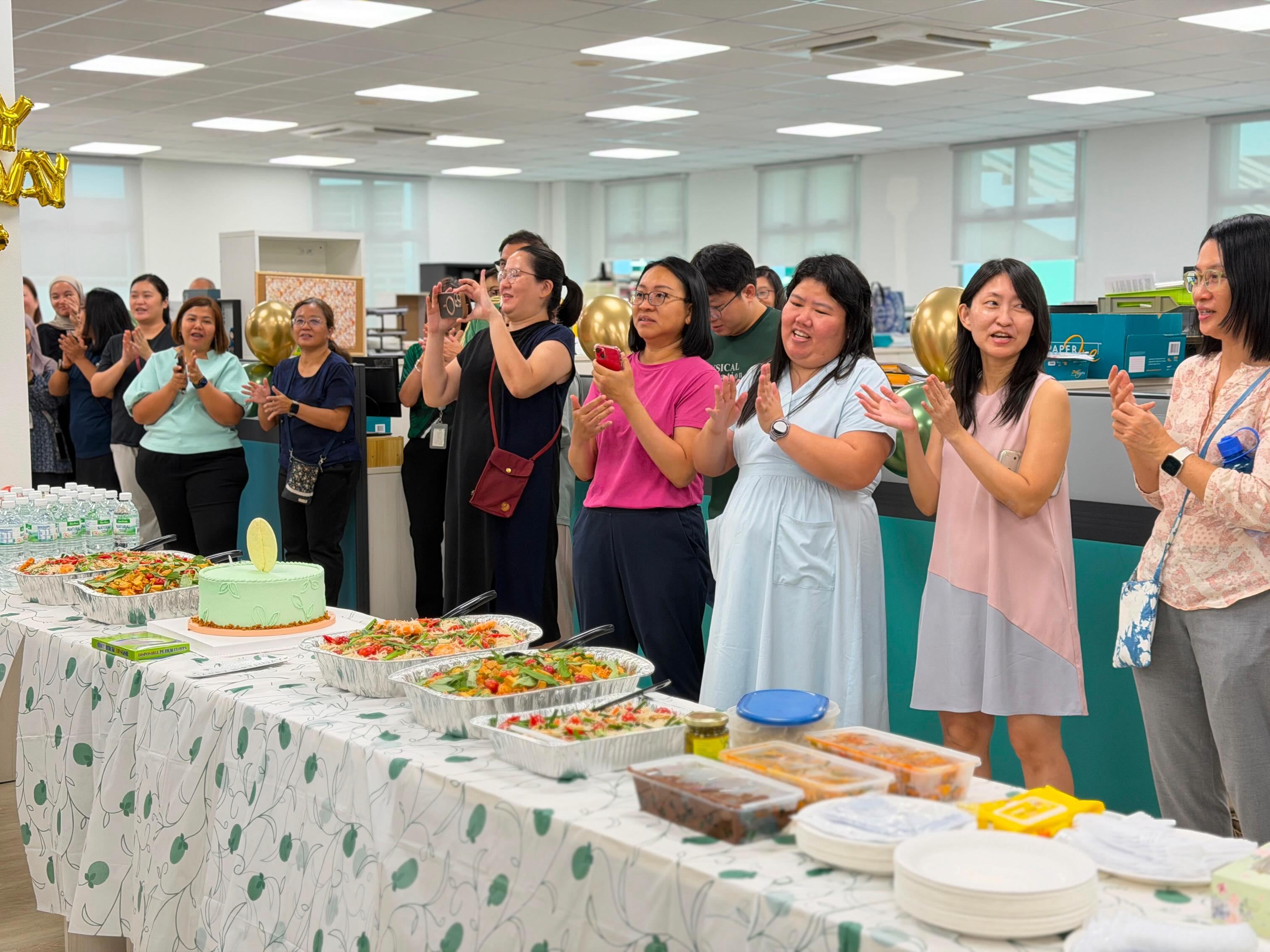 A large group of CTSS staff applauding and celebrating together around the cake and food spread in the newly renovated staffroom.