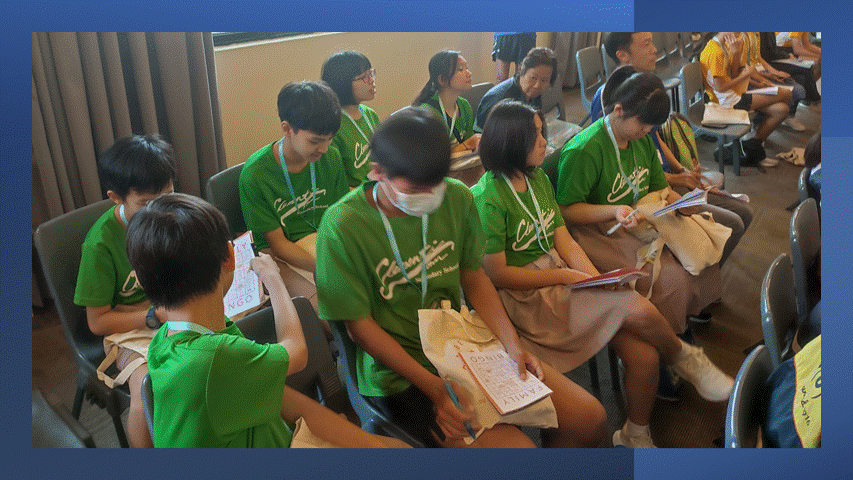 A group of students in green shirts sit in chairs holding papers and notebooks during a meeting or event.