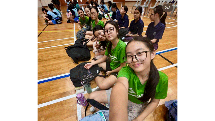 Several teenage girls in green shirts sit on a gymnasium floor taking a selfie, with more students seated behind them.