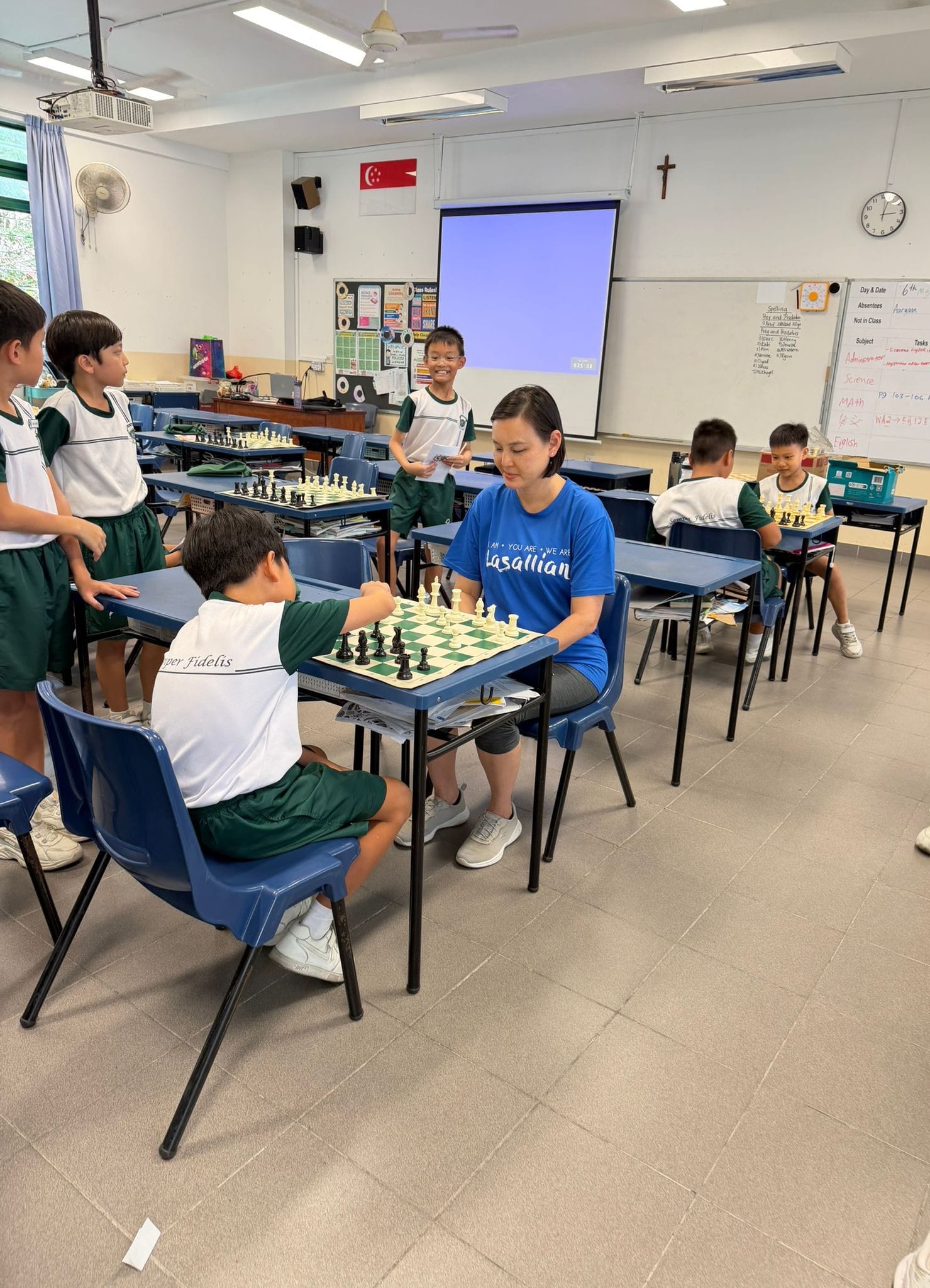 School Principal in a blue Lasallian t-shirt plays chess against a student in a classroom, while other students look on and play their own games in the background