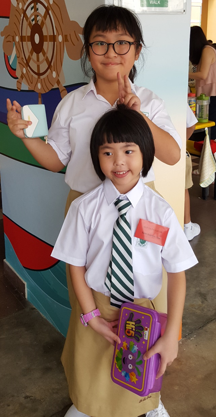 Two young girls in white school uniforms pose together, one holding a lunchbox and the other making a peace sign.