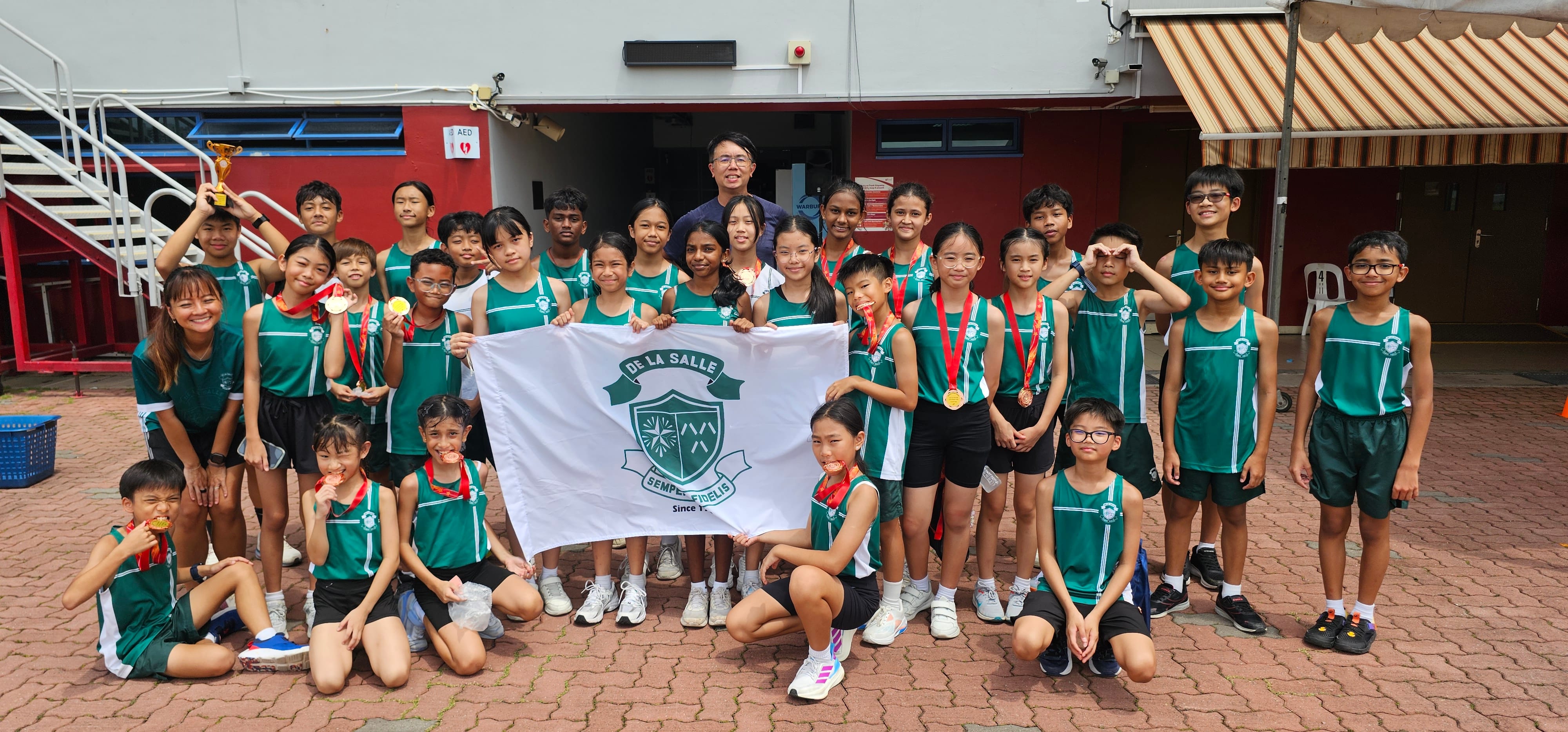 A proud group of students and a teacher posing together for a celebratory photo after a track and field competition, displaying their medals and trophies while holding up the school flag.