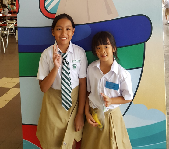 Two young girls in white shirts and khaki skirts pose smiling, one making a peace sign with her hand.