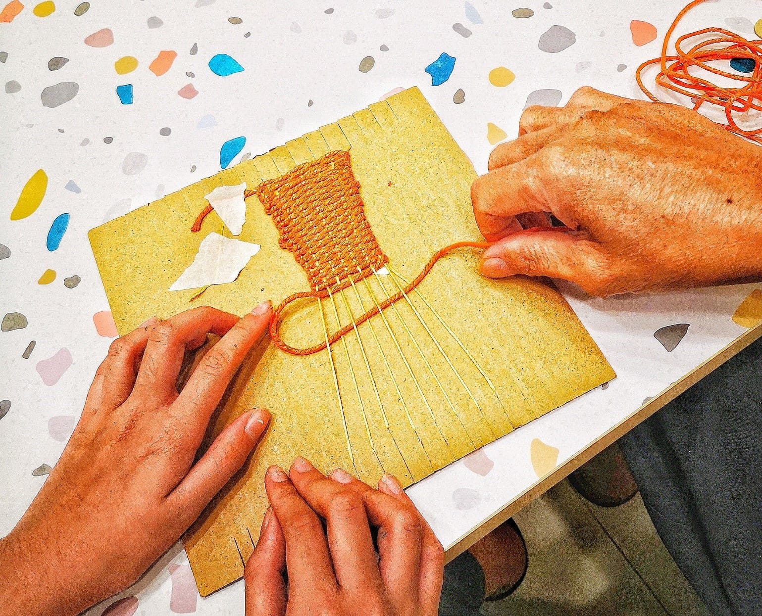 Two pairs of hands weaving orange yarn on a small cardboard loom placed on a colorful terrazzo table.