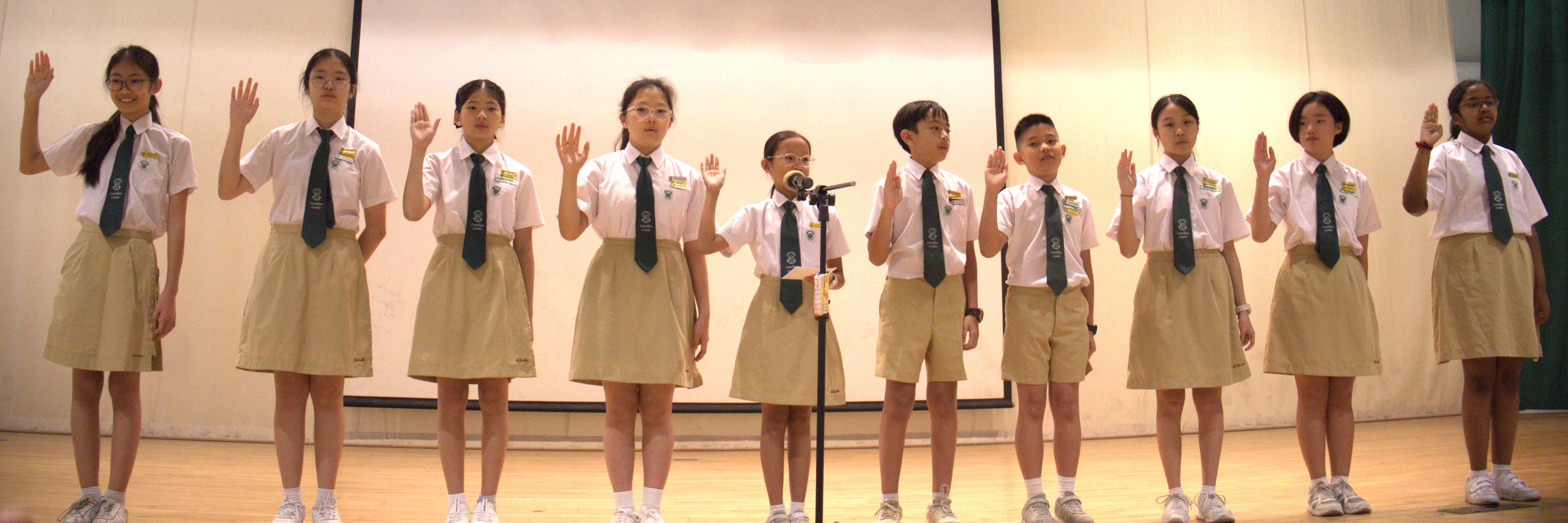 Student leaders in school uniform taking their pledge on stage during an investiture ceremony, with one student speaking at the microphone.