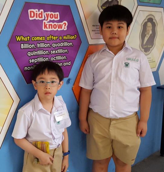 Two boys in school uniforms stand before a colorful "Did you know?" math facts display board.