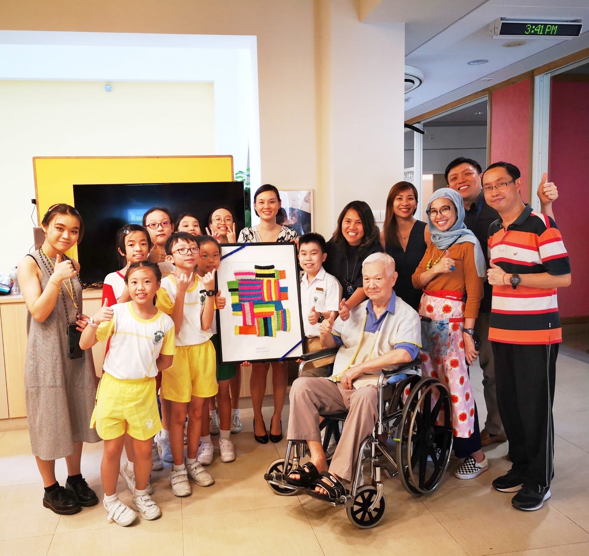 A group of children and adults pose with an elderly man in a wheelchair, holding a colorful framed artwork indoors.
