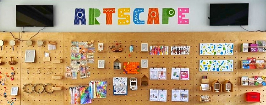 Colorful "ARTSCAPE" letters above a pegboard wall displaying children's artwork, crafts, and small wooden objects.