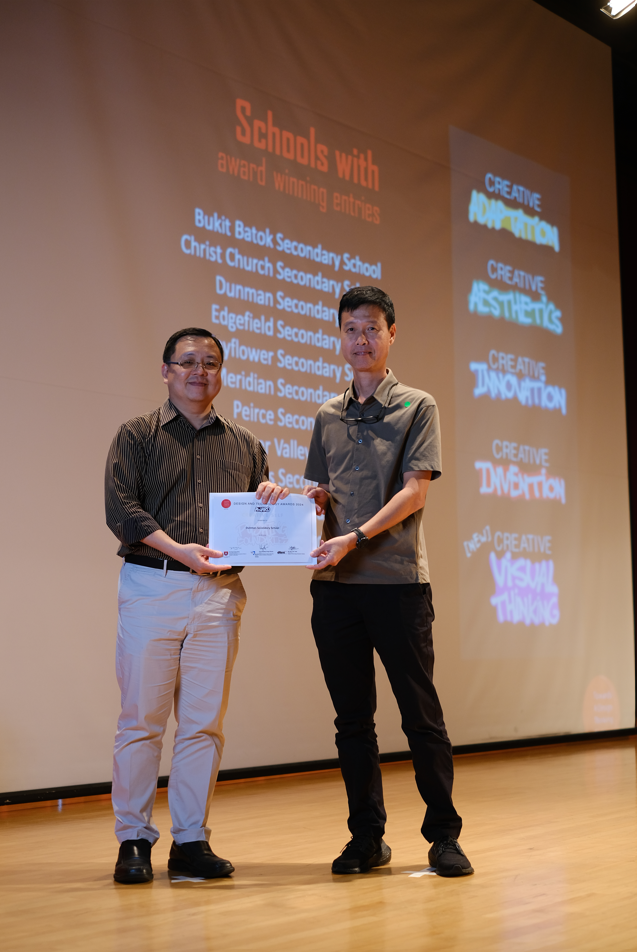 Two men stand on a stage holding a certificate, with a projected screen listing schools with award winning entries behind them.