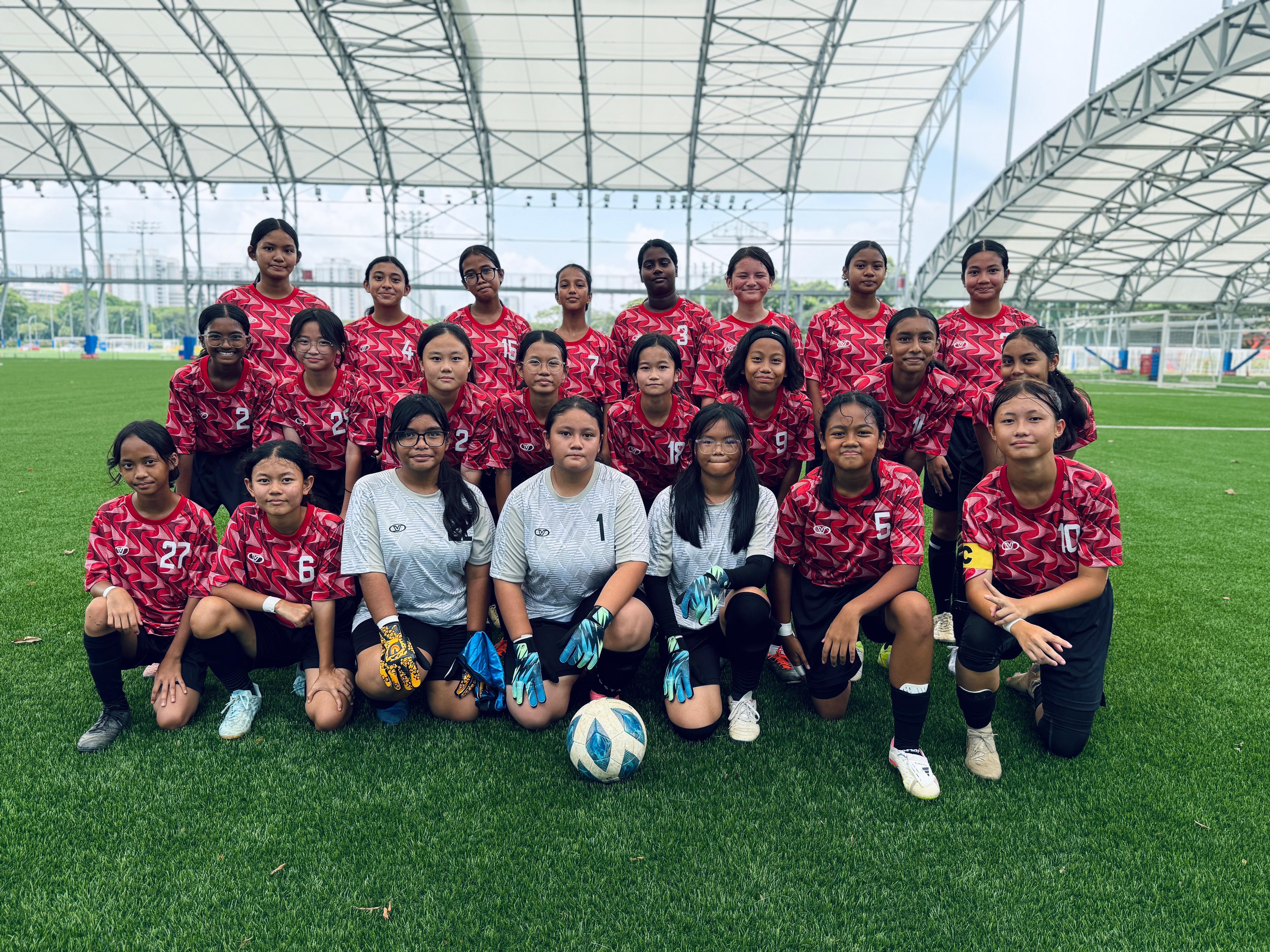 A girls football team in red uniforms poses on a green turf field under a covered stadium roof.