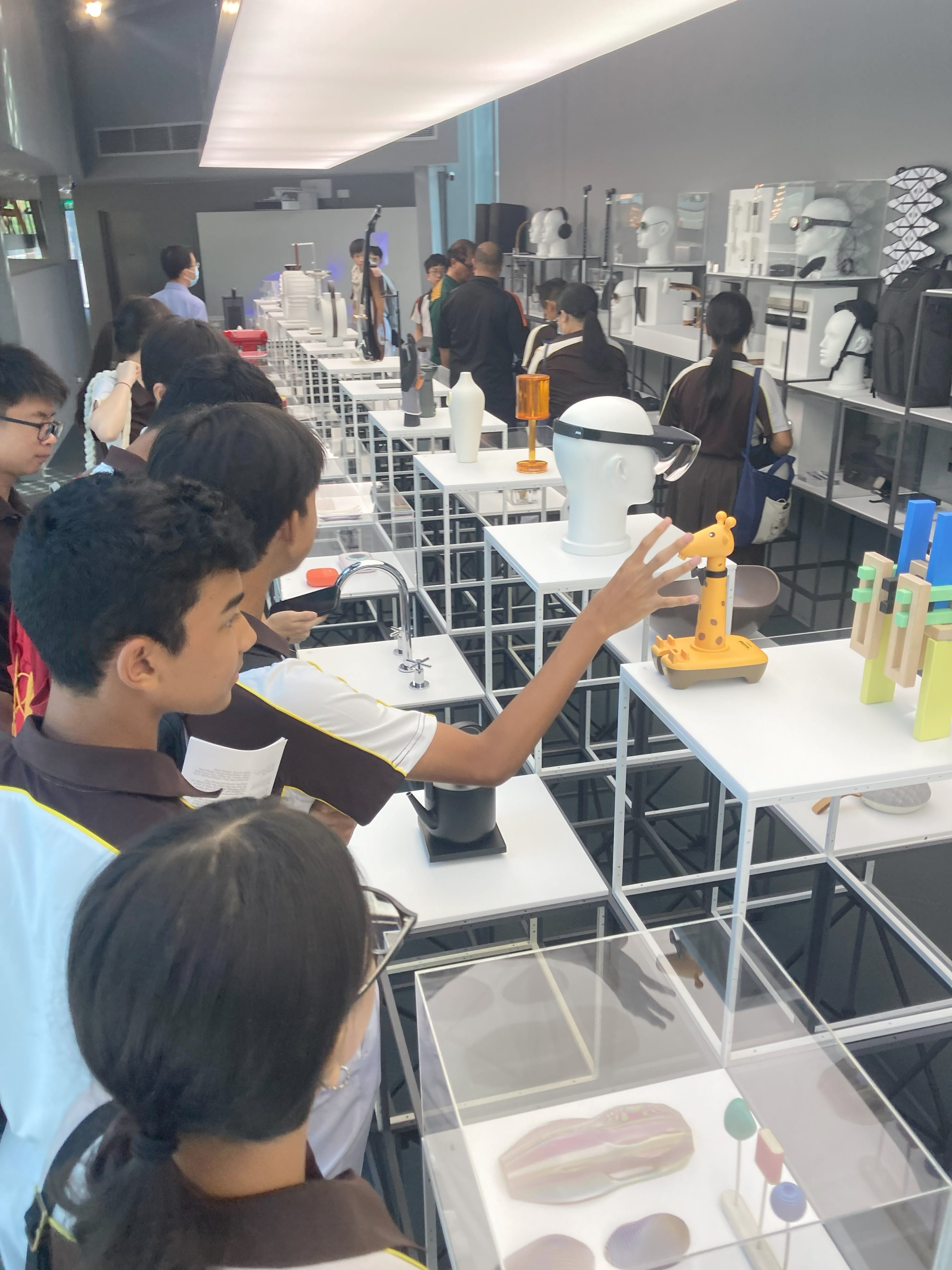 Students browse a design exhibition with white display tables holding objects like a giraffe toy, mannequin head, and faucet.