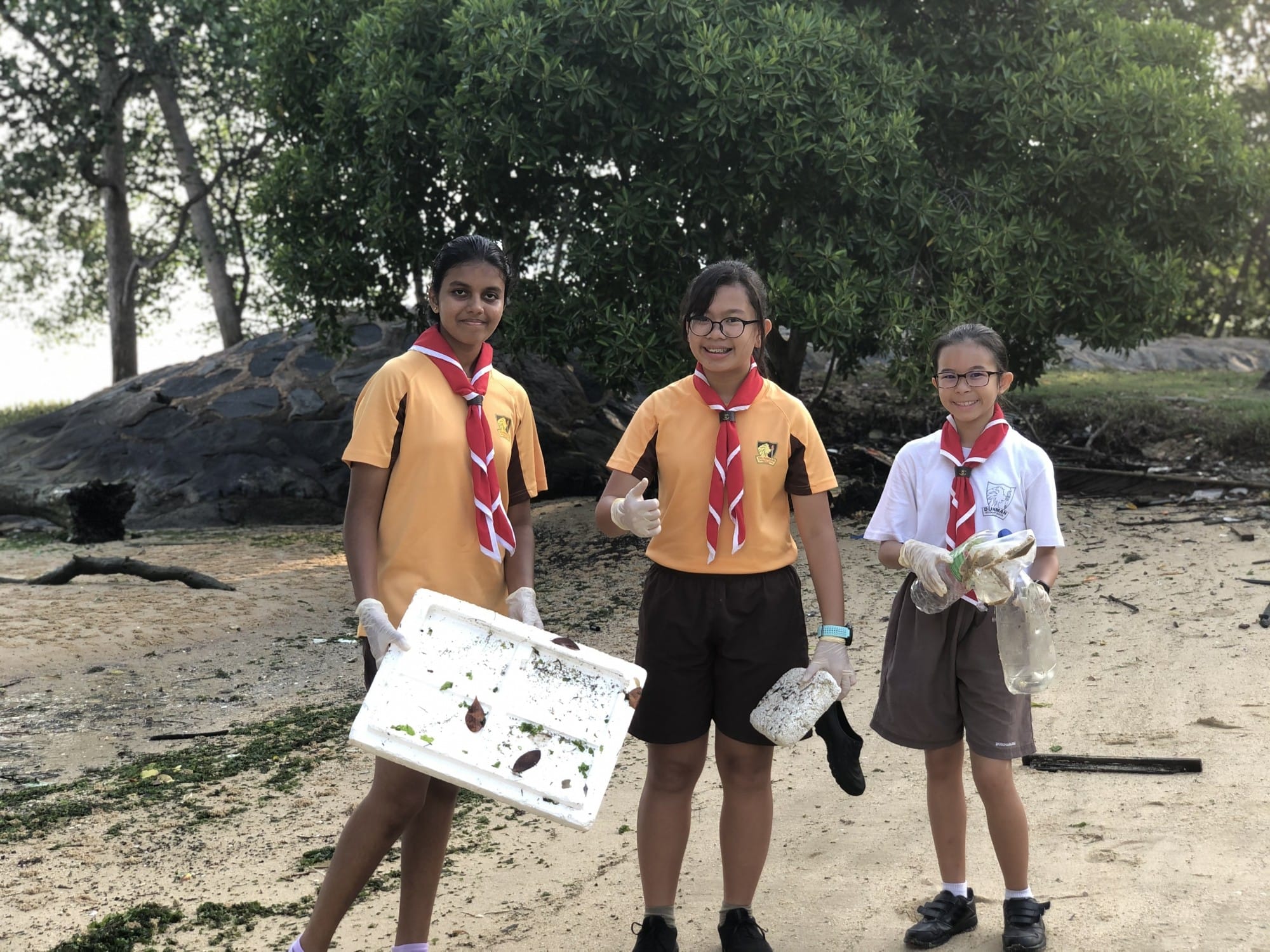 Three young students in school uniforms and red scarves hold collected trash while cleaning a beach near trees.