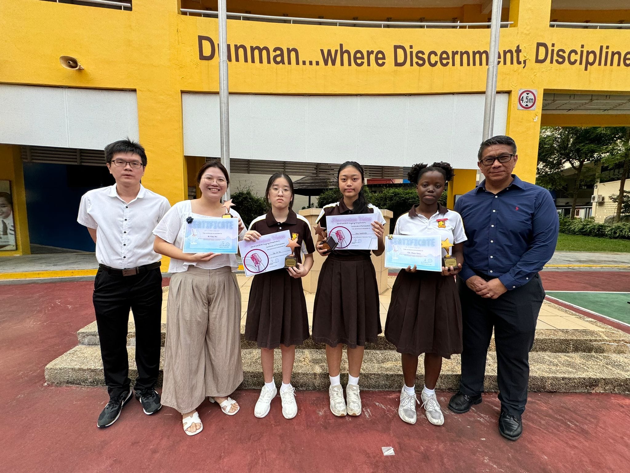 Six people pose outdoors at Dunman school, holding certificates and trophies in front of a yellow building.