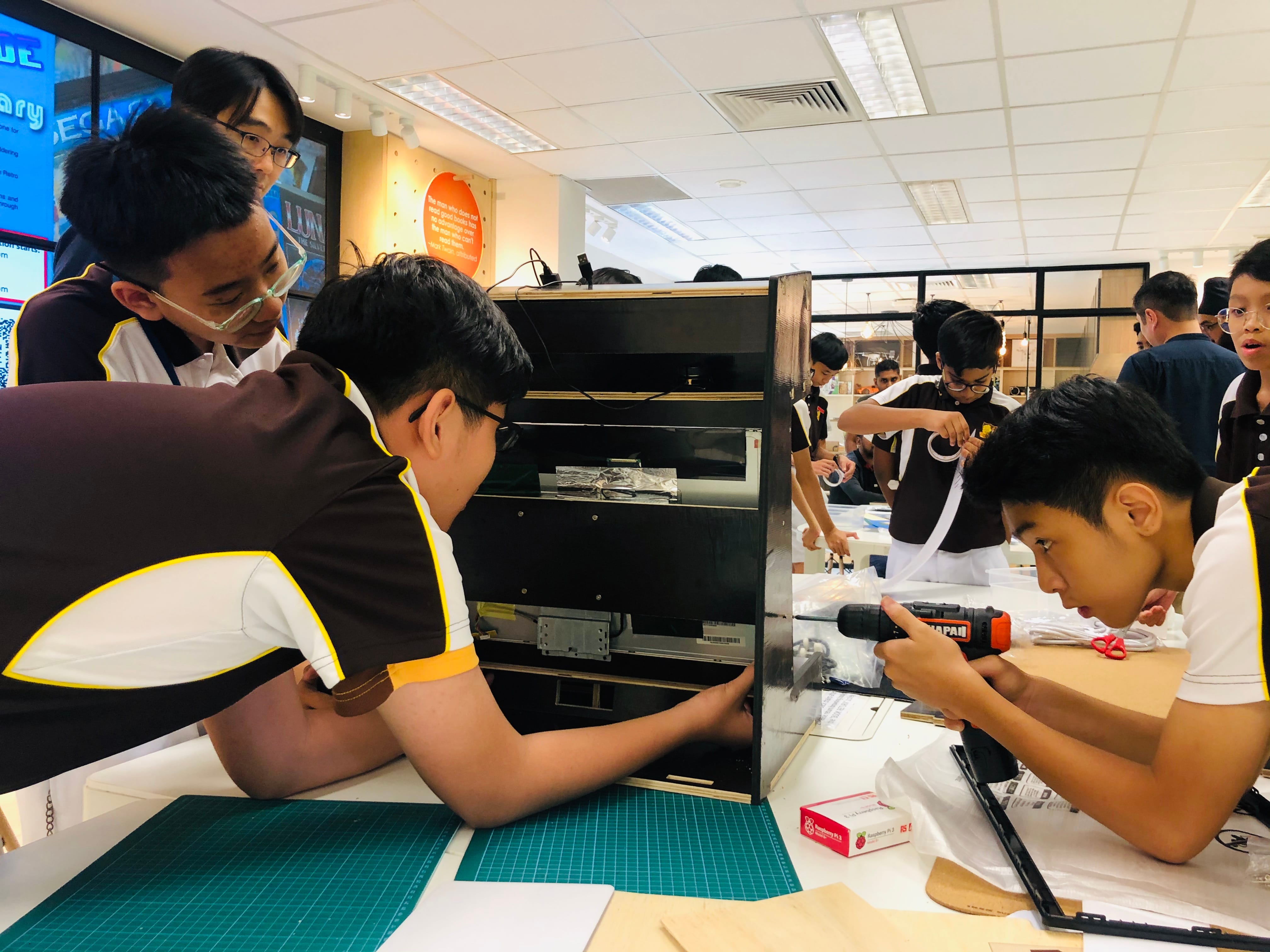 Students in brown uniforms assemble a wooden cabinet using a power drill in a classroom workshop.