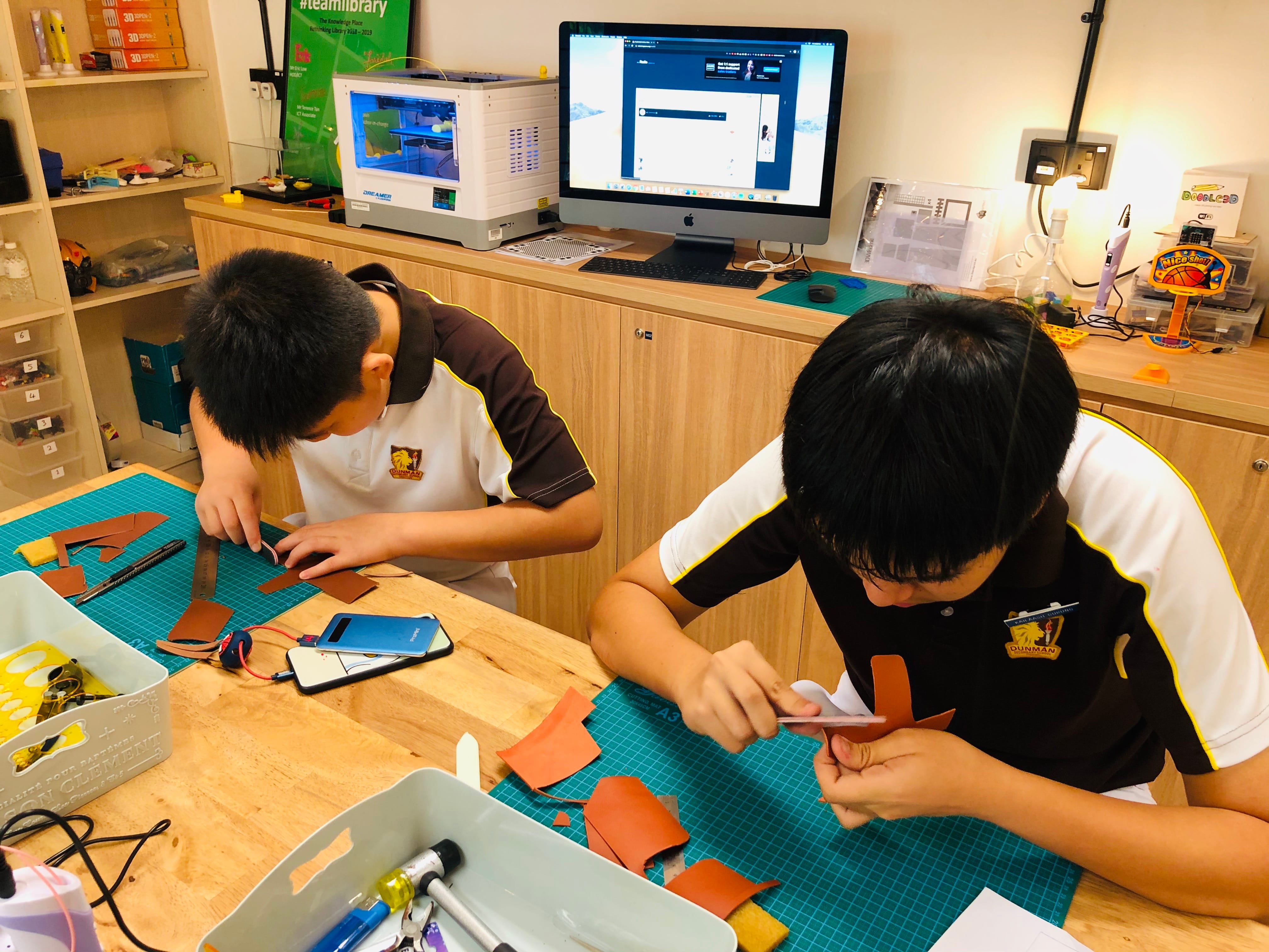 Two students in school uniforms work with leather pieces on a craft mat in a makerspace with a 3D printer nearby.