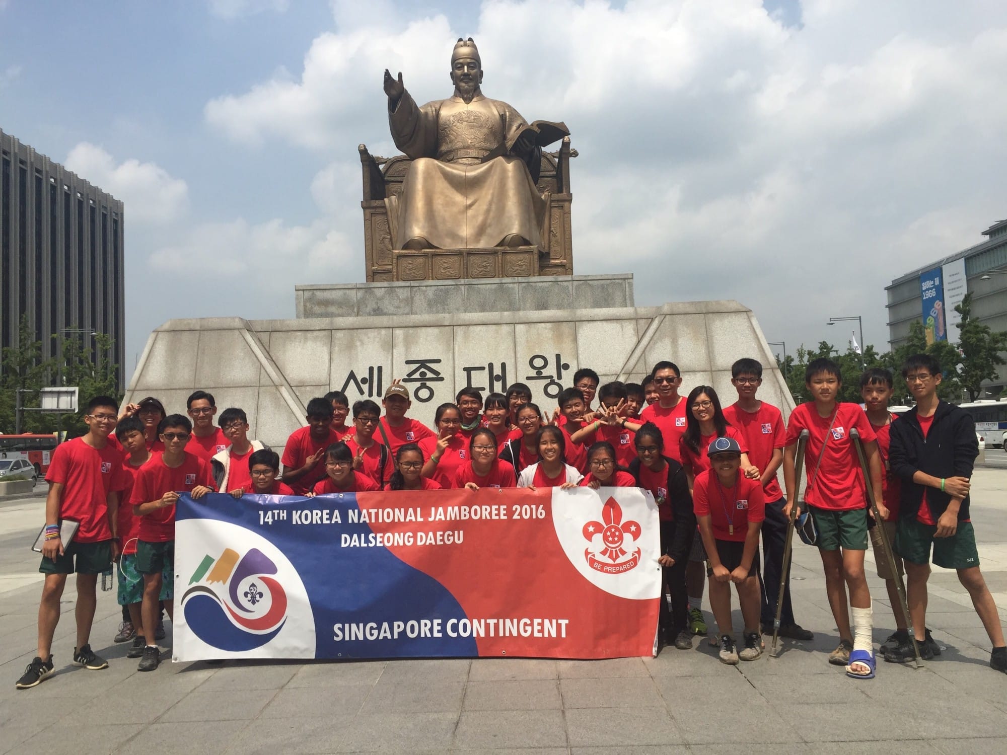 Group in red shirts holds a Singapore Contingent banner for the 14th Korea National Jamboree 2016 before a seated bronze statue.