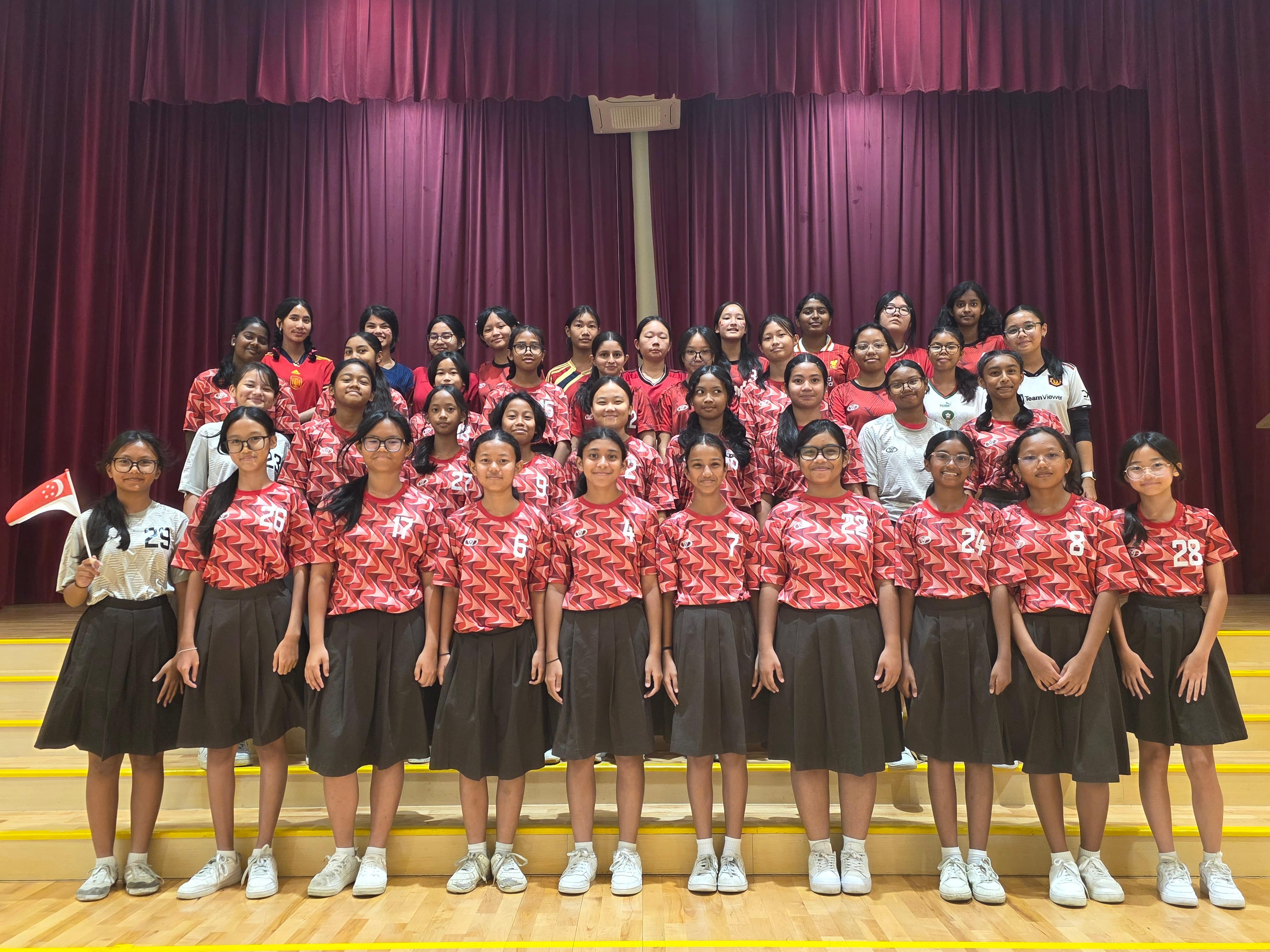 A large group of girls in numbered red jerseys and dark skirts pose on stage steps before a red curtain.