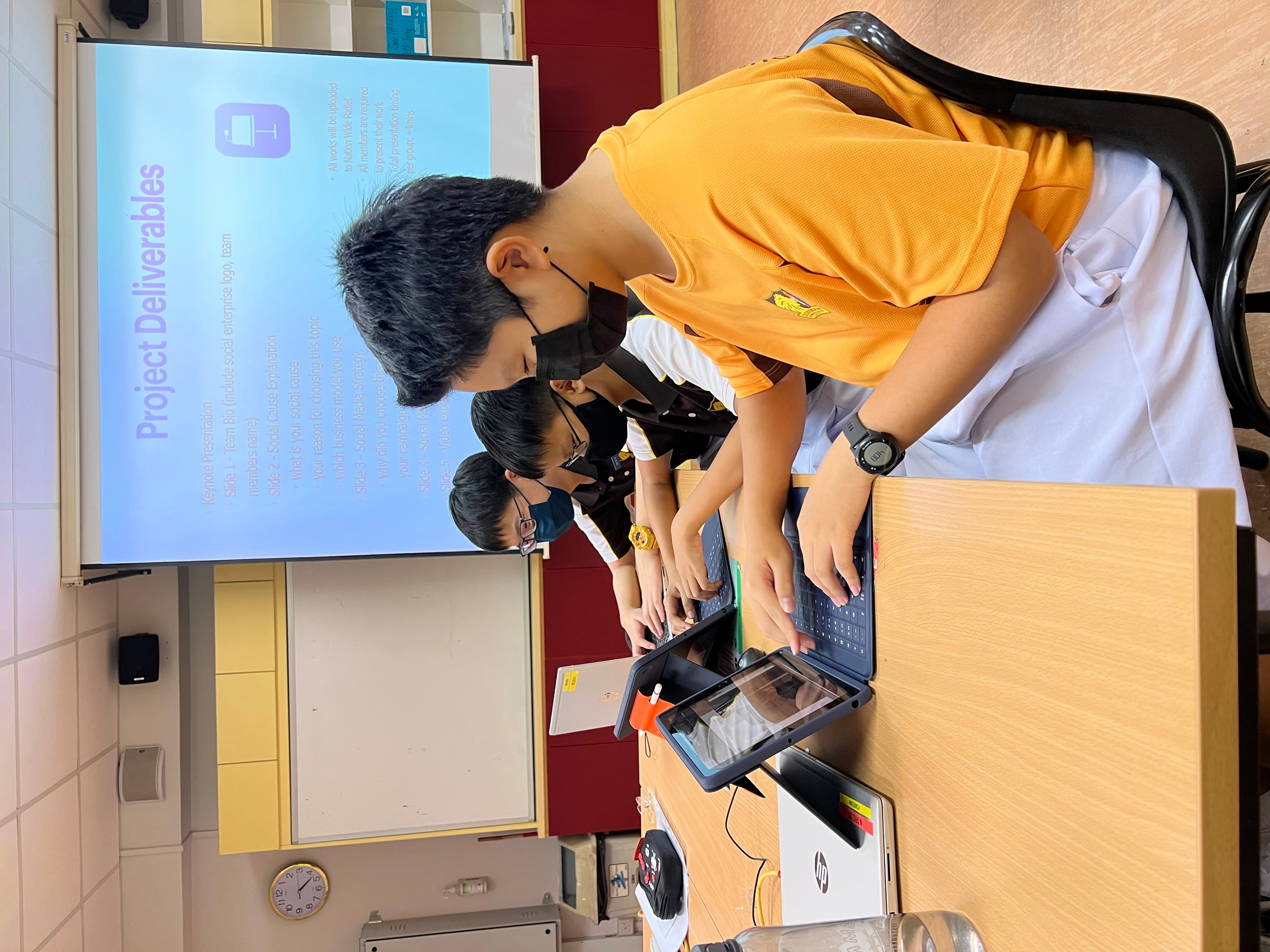 Three masked students work on laptops at a desk facing a projected slide titled Project Deliverables.