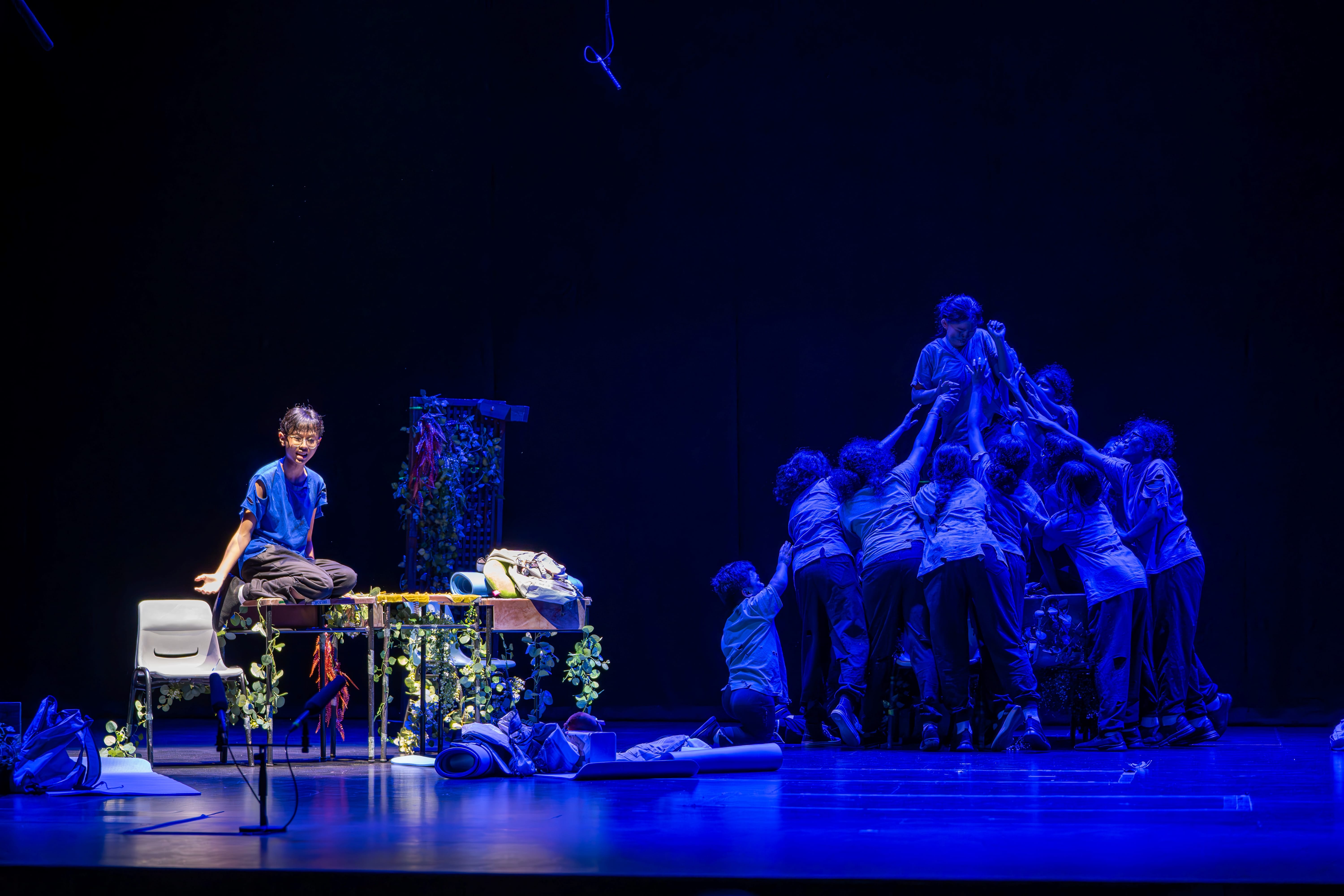 Blue-lit stage: one person sits by a cluttered table, while a group lifts someone in a tight huddle.